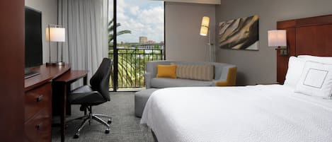 A hotel room with a bed, a desk, a chair, a sofa, a balcony with a view, and a painting on the wall.