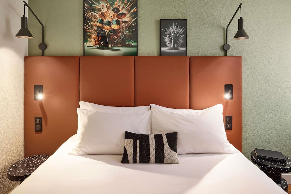 A hotel room with a large bed, two wall-mounted reading lamps, a framed artwork above the bed, and a small side table with a book.