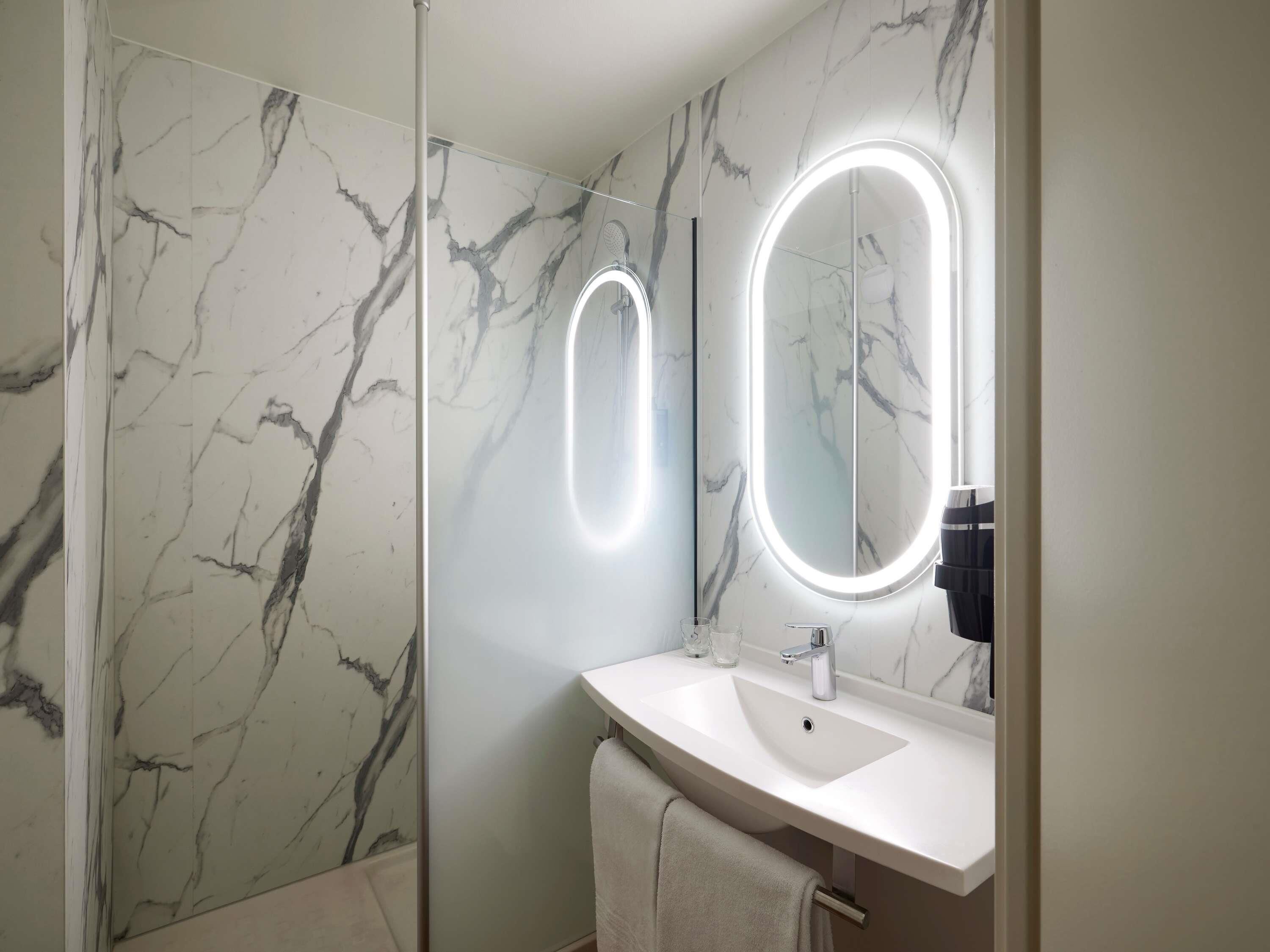 A bathroom with marble walls, a white sink, and two illuminated mirrors.