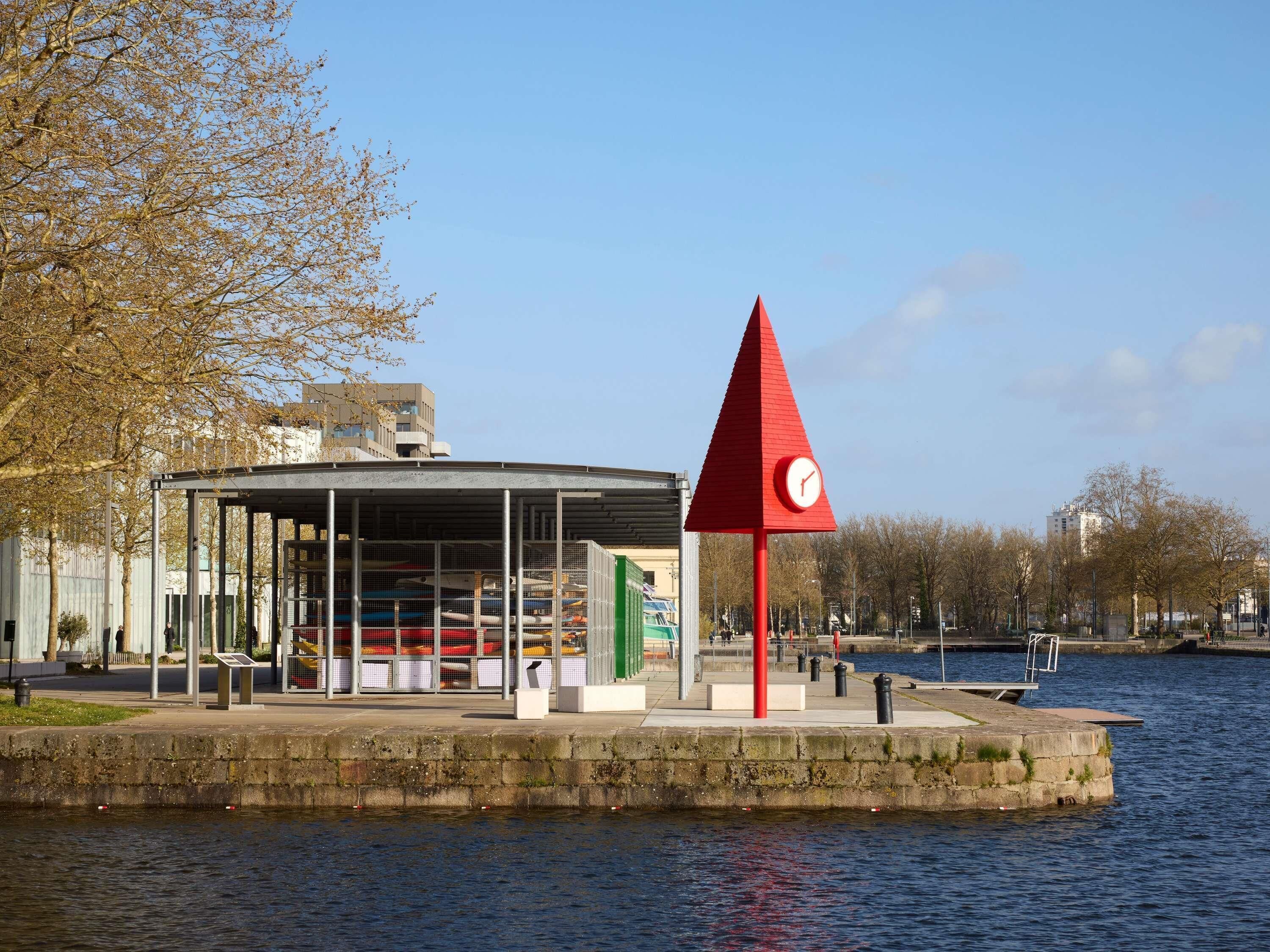 A waterfront building with a red clock tower, a covered area with seating, and a body of water.