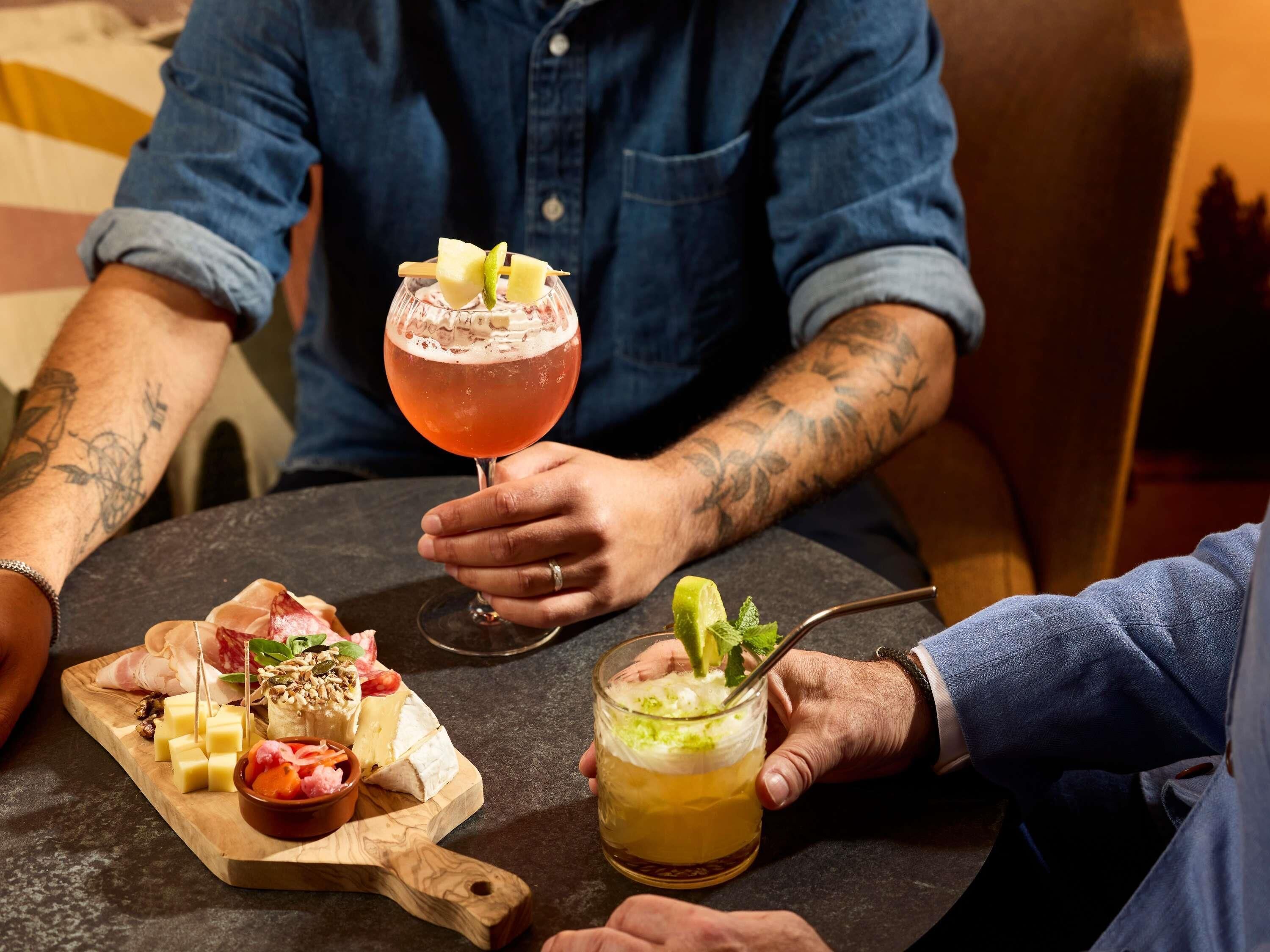 Une table ronde avec une planche de fromages, un cocktail et une personne tenant un verre.