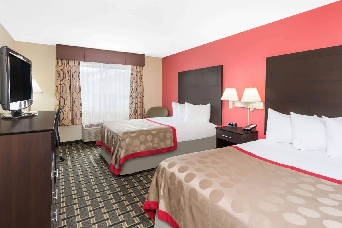 A hotel room with two beds, a desk with a TV, a window with curtains, and a red accent wall.