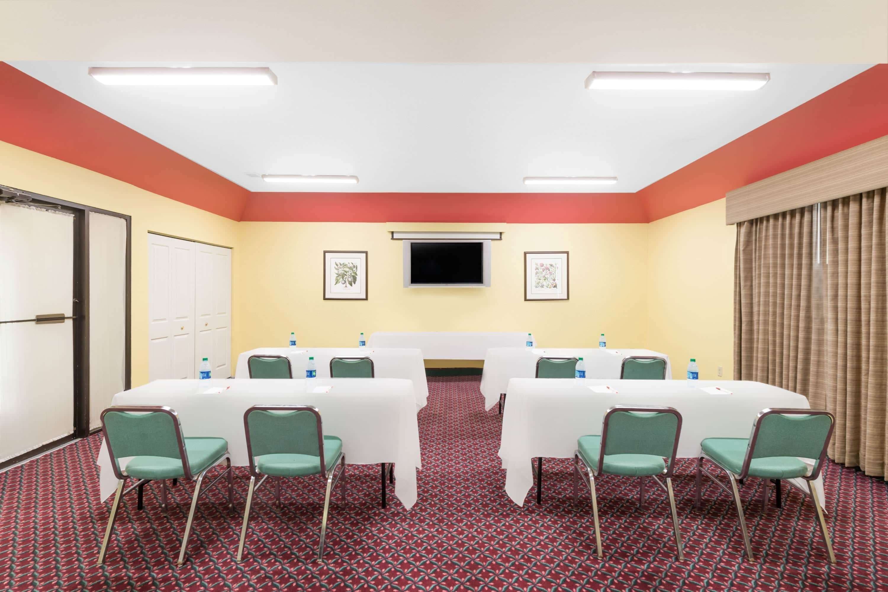 A conference room with a long table, green chairs, a flat-screen TV, framed pictures, and a red carpet.