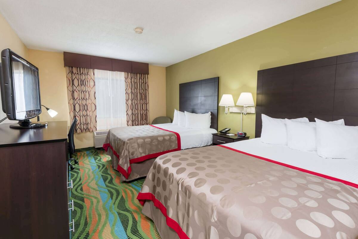 A hotel room with two beds, a desk with a television, a window with patterned curtains, and a patterned carpet.
