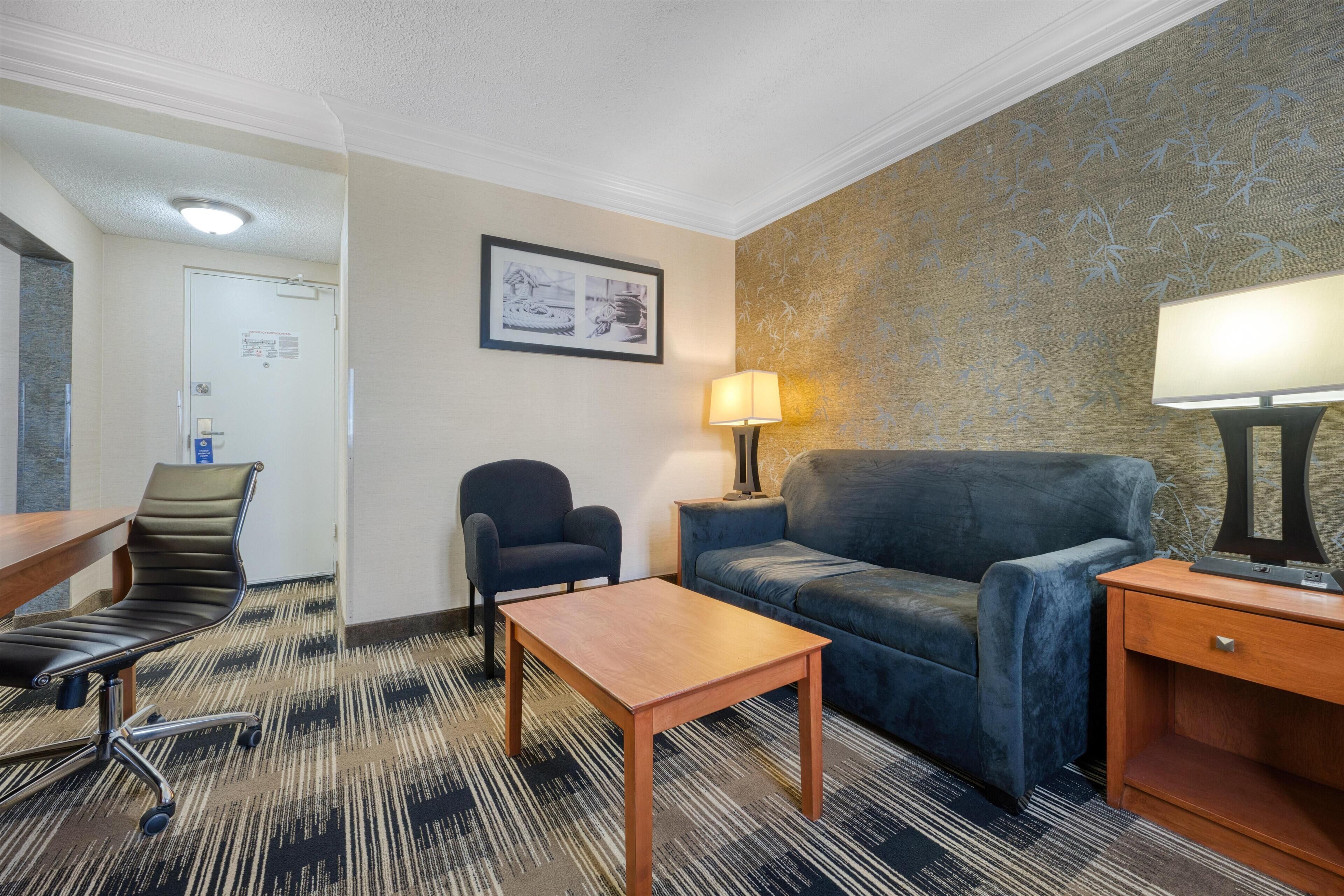 Une chambre d’hôtel avec un canapé, un fauteuil, une petite table, une lampe et un sol recouvert de moquette.