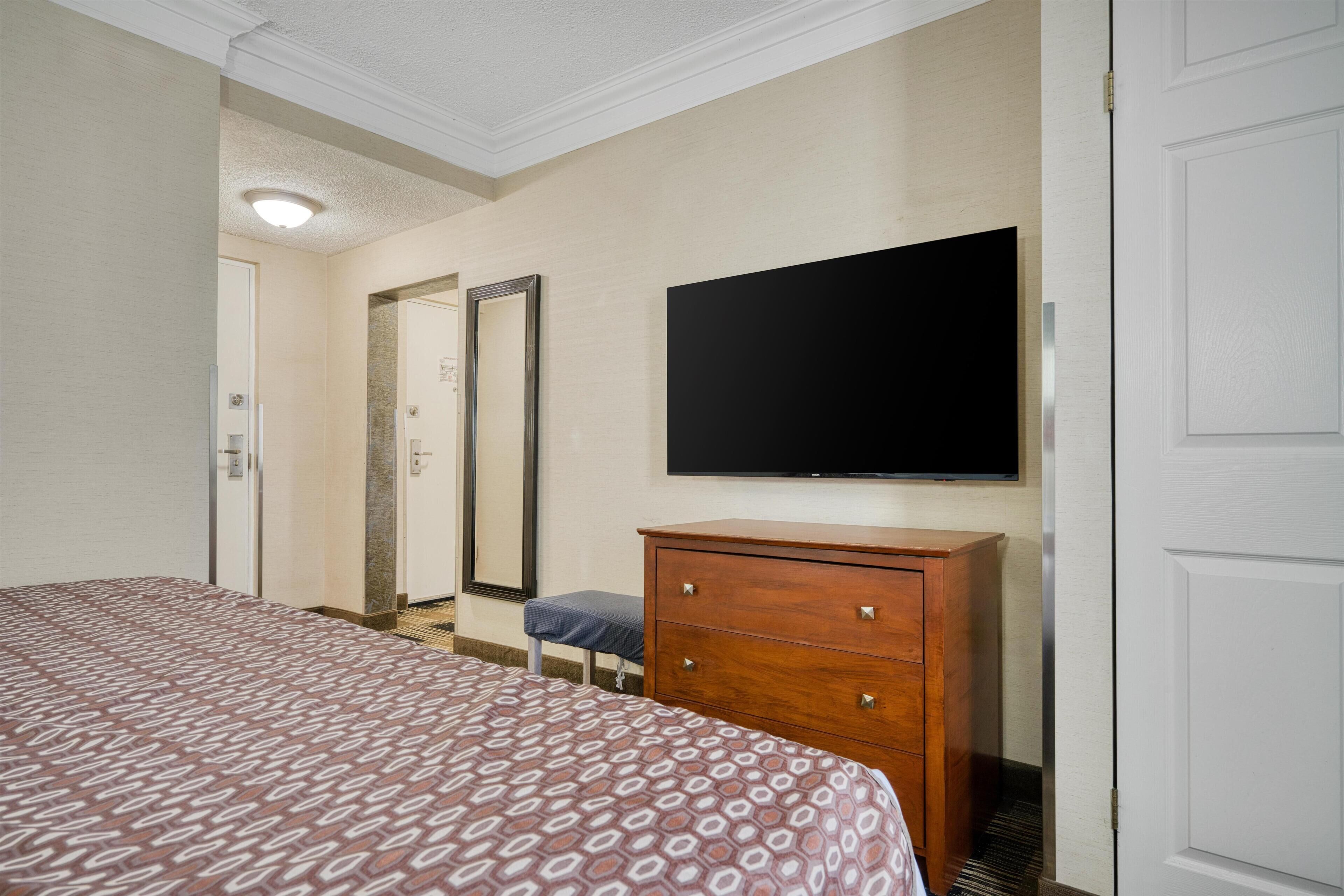 A hotel room with a bed, a wooden dresser, a flat-screen TV mounted on the wall, a mirror, and a door.