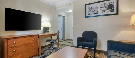 Una camera d'albergo con TV a schermo piatto, comò in legno, scrivania, sedia, divano e un quadro appeso al muro.