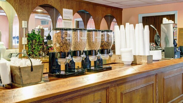 A breakfast bar with multiple dispensers for cereal and granola, a coffee machine, and stacks of white paper cups. - Rodeway Inn New Braunfels - San Antonio Area (New Braunfels)