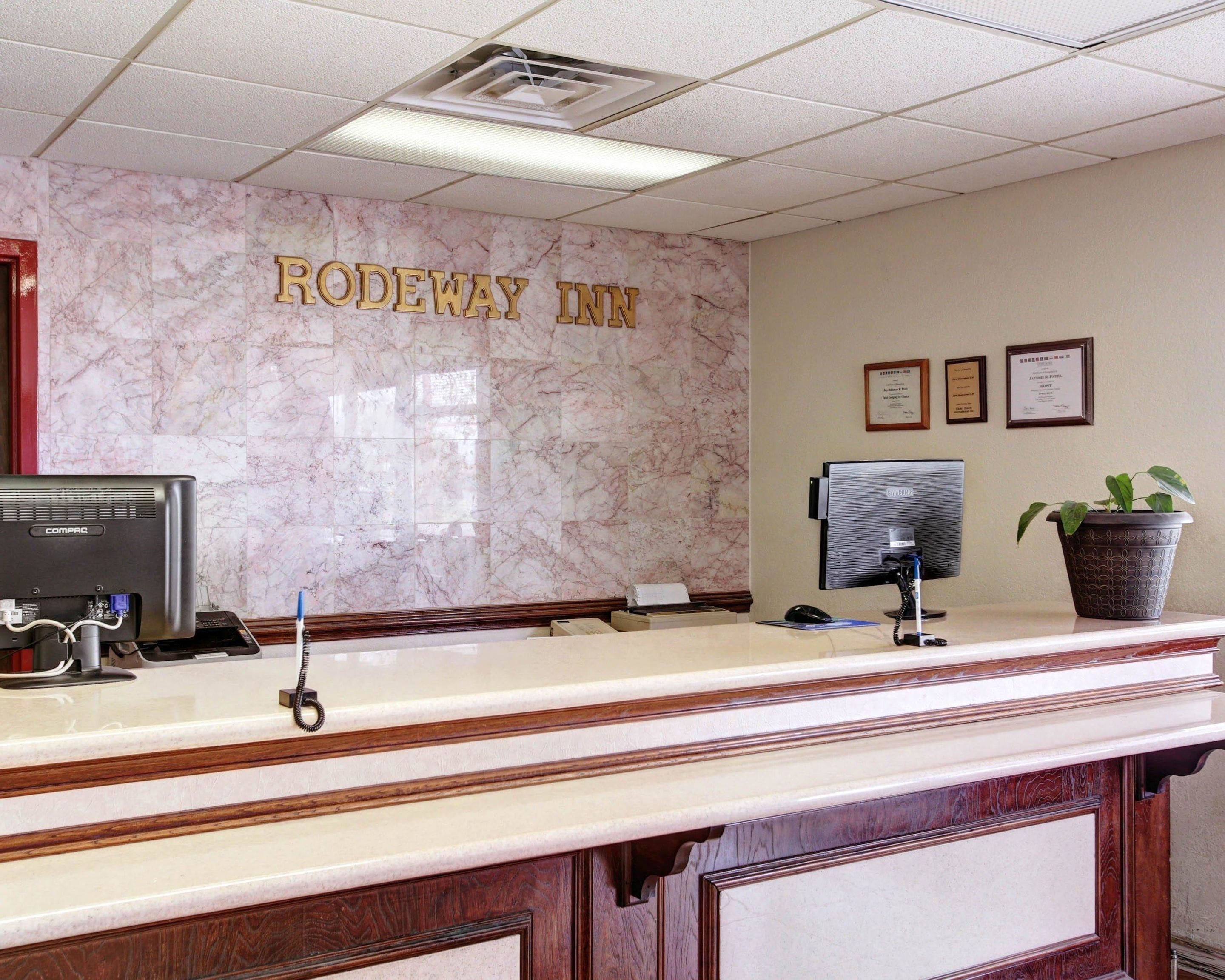 A reception desk at Rodeway Inn with a marble wall, a computer, a phone, and a potted plant.