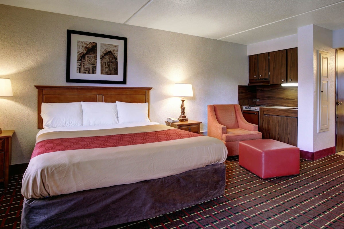 A hotel room with a large bed, a chair, a small table, a lamp, a framed picture, and a kitchenette.