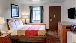 A hotel room with a bed, bedside table, lamp, window with curtains, door, and a television on the wall. - Rodeway Inn New Braunfels - San Antonio Area (New Braunfels)