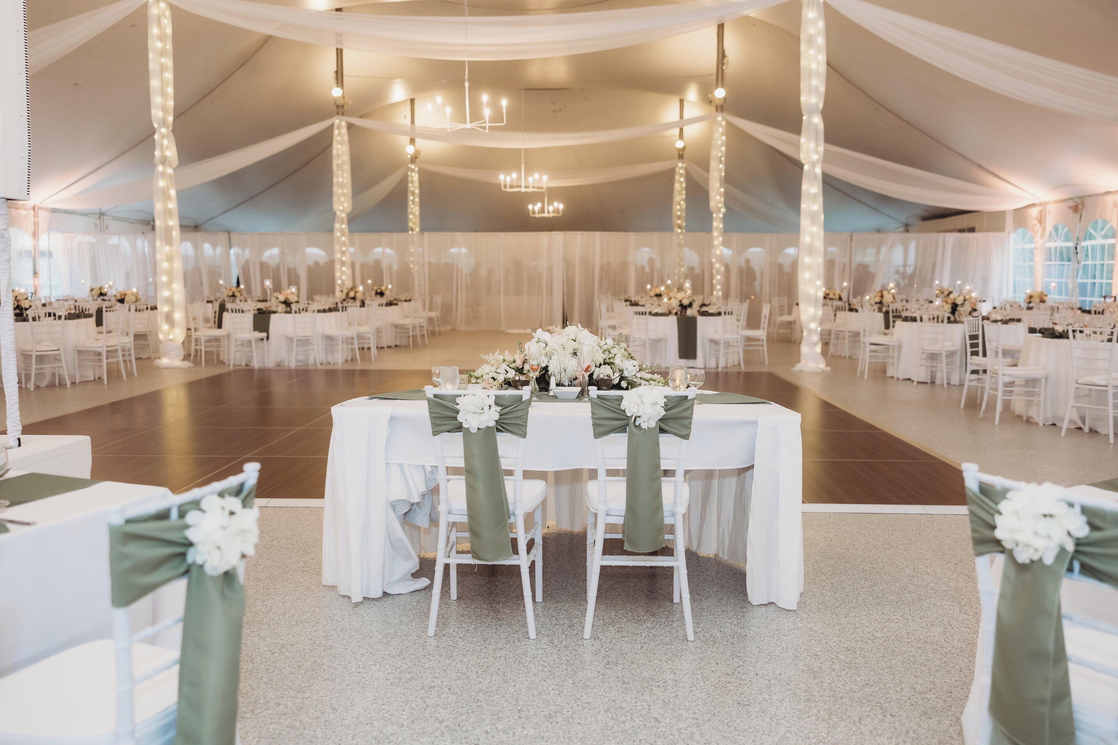 A tented wedding venue with white tables and chairs, chandeliers, and floral centerpieces.