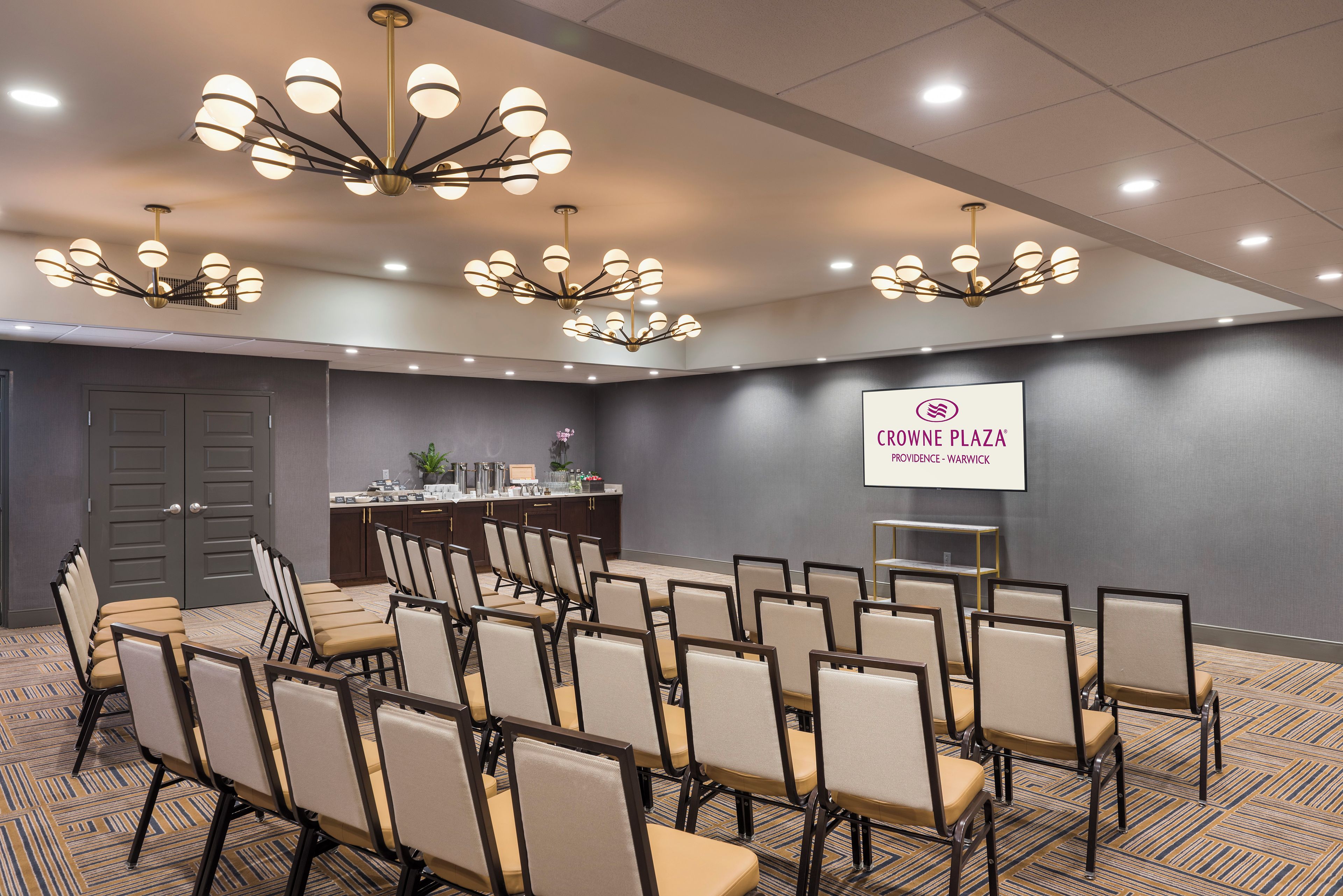 A conference room with rows of chairs, a podium, and a screen displaying the logo 'CROWNE PLAZA PROVIDENCE-WARWICK'.