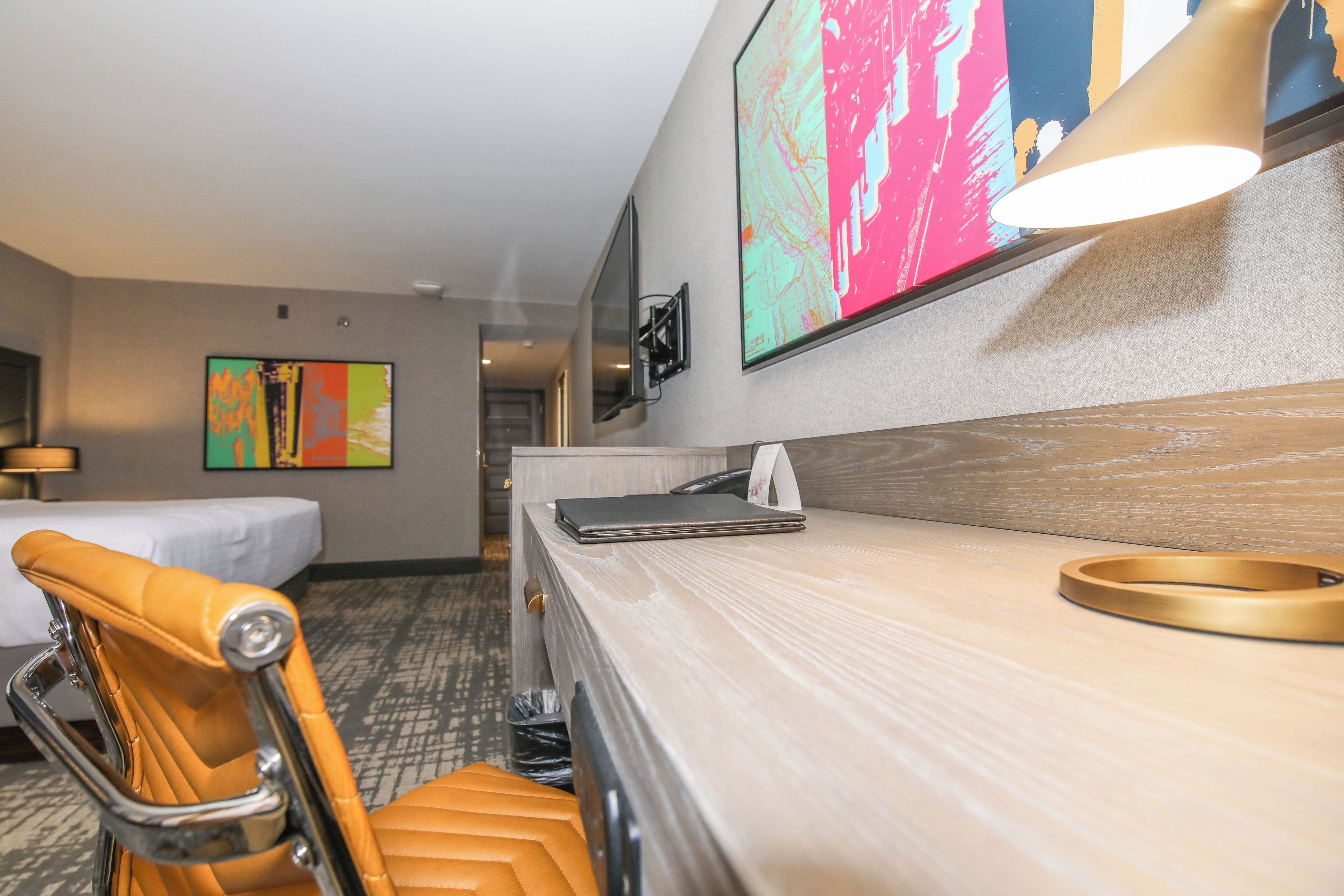 A hotel room with a bed, a desk with a lamp, a chair, and a large abstract painting on the wall.