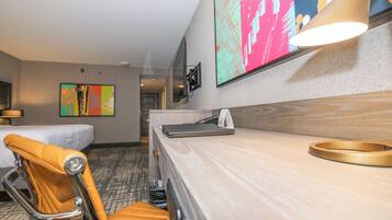 A hotel room with a bed, a desk with a lamp, a chair, and a large abstract painting on the wall.