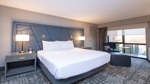 Une chambre d’hôtel moderne avec un grand lit, deux tables de chevet avec des lampes, un petit canapé et une grande fenêtre donnant sur la ville.