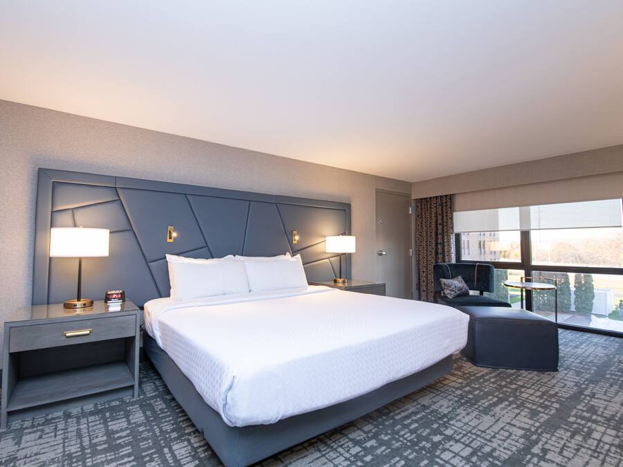 A modern hotel room with a large bed, two bedside tables with lamps, a small sofa, and a large window offering a city view.