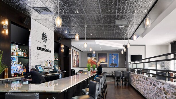 Uma área de bar com um balcão comprido, banquetas e uma parede de tijolos. Há luminárias pendentes e uma placa com a inscrição 'The Crossings'.