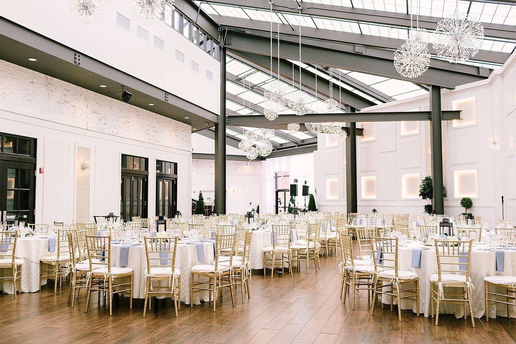 Un vaste espace intérieur doté de hauts plafonds, de nombreuses tables recouvertes de nappes blanches et entourées de chaises dorées, ainsi que de luminaires décoratifs suspendus.