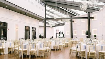 Un vaste espace intérieur doté de hauts plafonds, de nombreuses tables recouvertes de nappes blanches et entourées de chaises dorées, ainsi que de luminaires décoratifs suspendus.