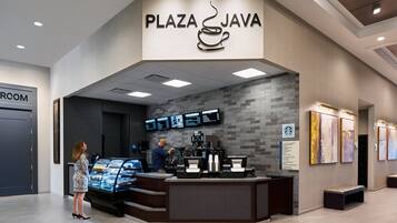A modern coffee shop named 'Plaza Java' with a digital menu display, a coffee machine, and a glass display case of pastries.