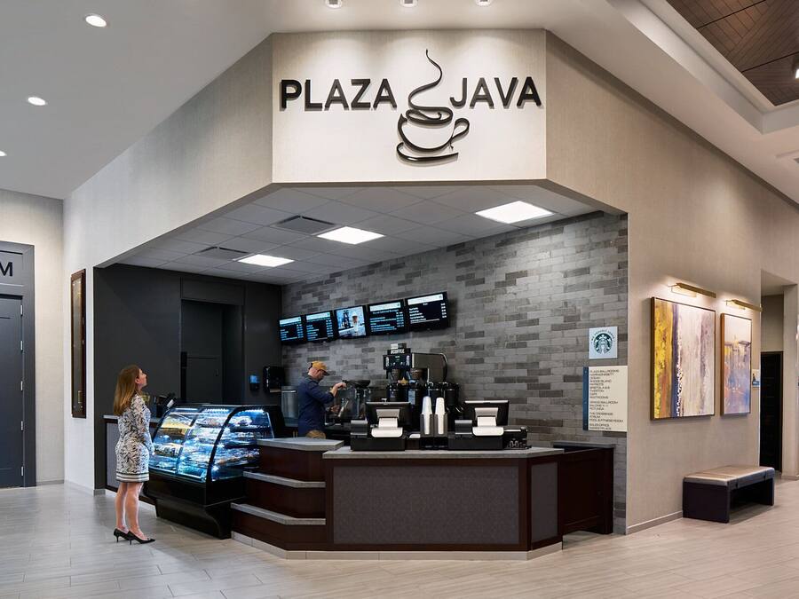 A modern coffee shop named 'Plaza Java' with a digital menu display, a coffee machine, and a glass display case of pastries.