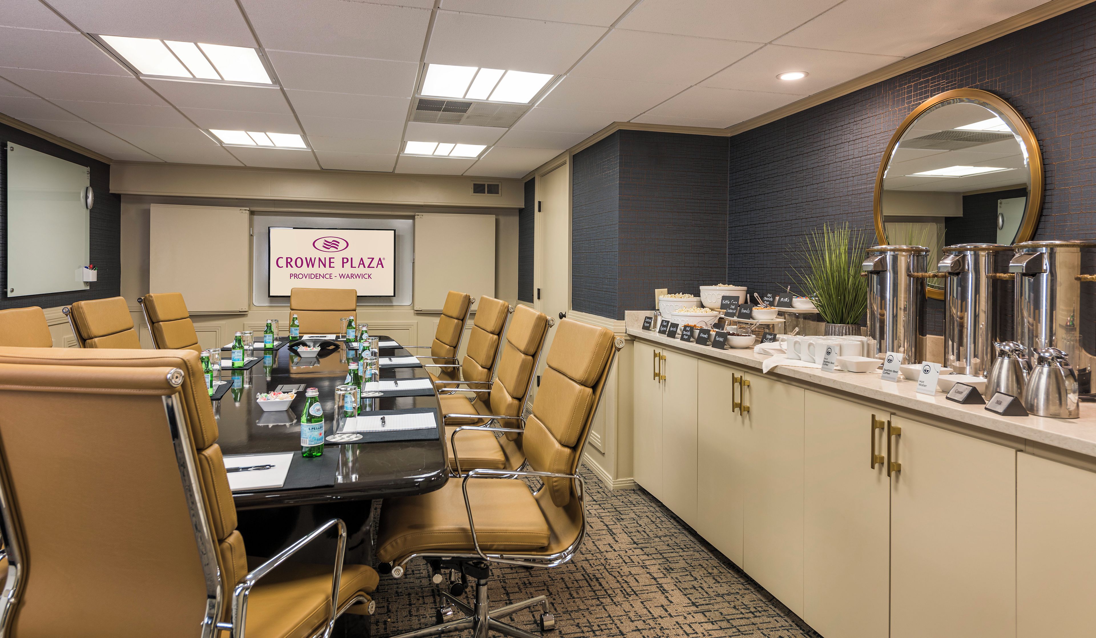 A conference room with a long table, chairs, a projector screen, and a buffet area with food and drink options.