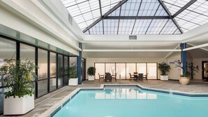 Une piscine intérieure dotée d’un toit en verre, entourée de plantes en pot et d’espaces aménagés pour s’asseoir.