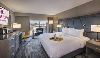 Habitación de hotel con una cama grande, mesitas de noche, un escritorio con cafetera, una silla y vistas al exterior.