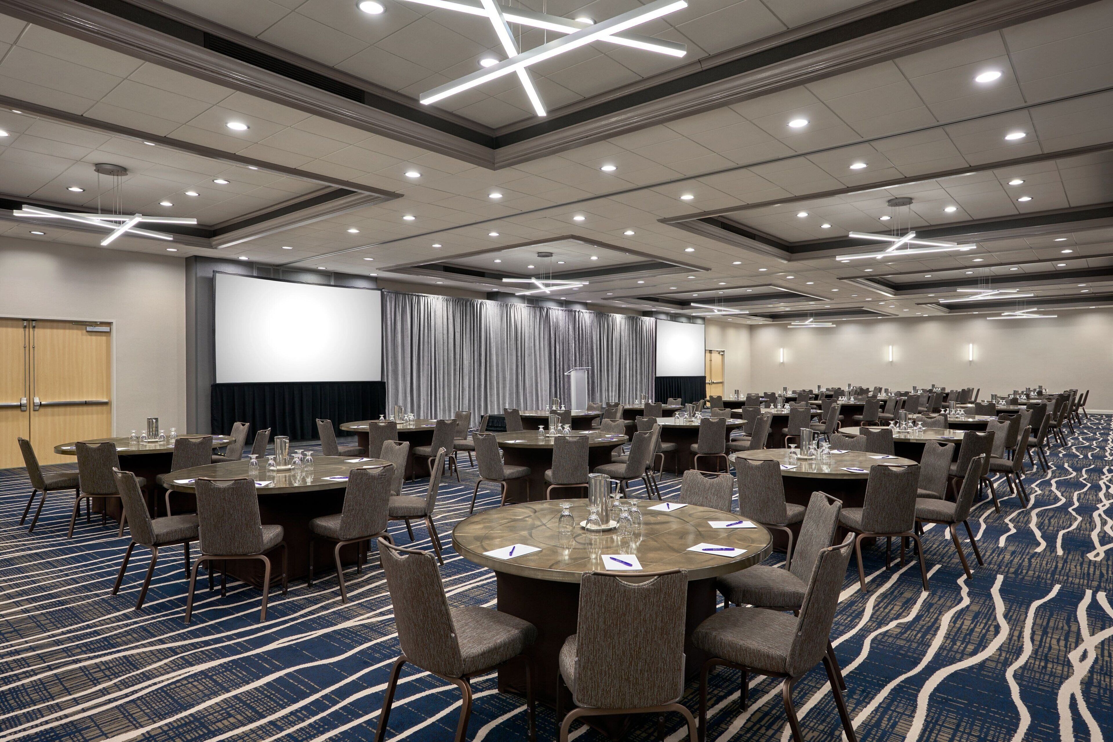 A conference room with multiple tables and chairs, a large screen, and a carpeted floor.