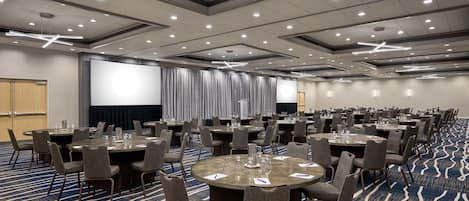 Un salón de conferencias con varias mesas y sillas, una pantalla grande y suelo alfombrado.