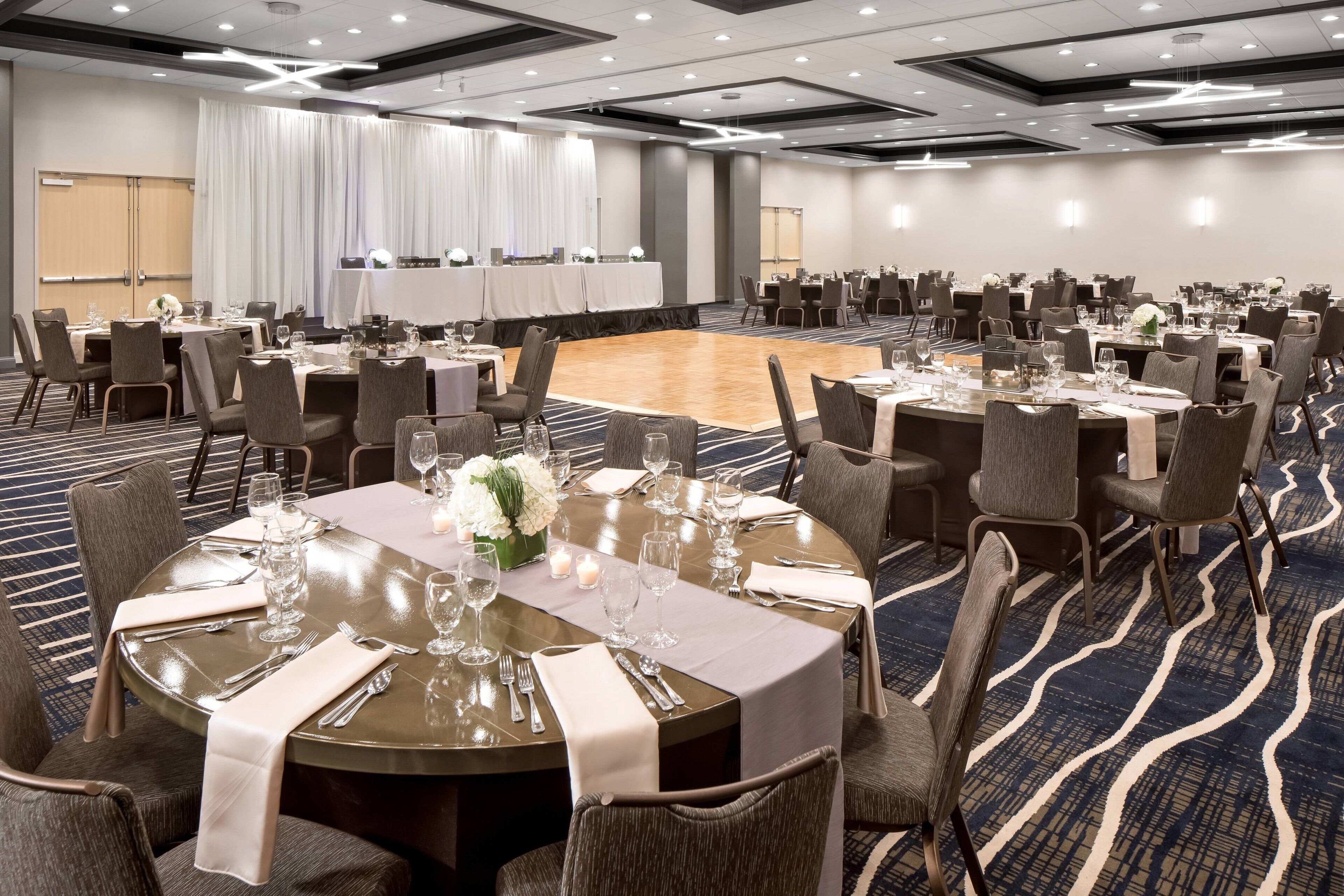 A banquet hall with round tables set for dining, a dance floor, and a stage with curtains.