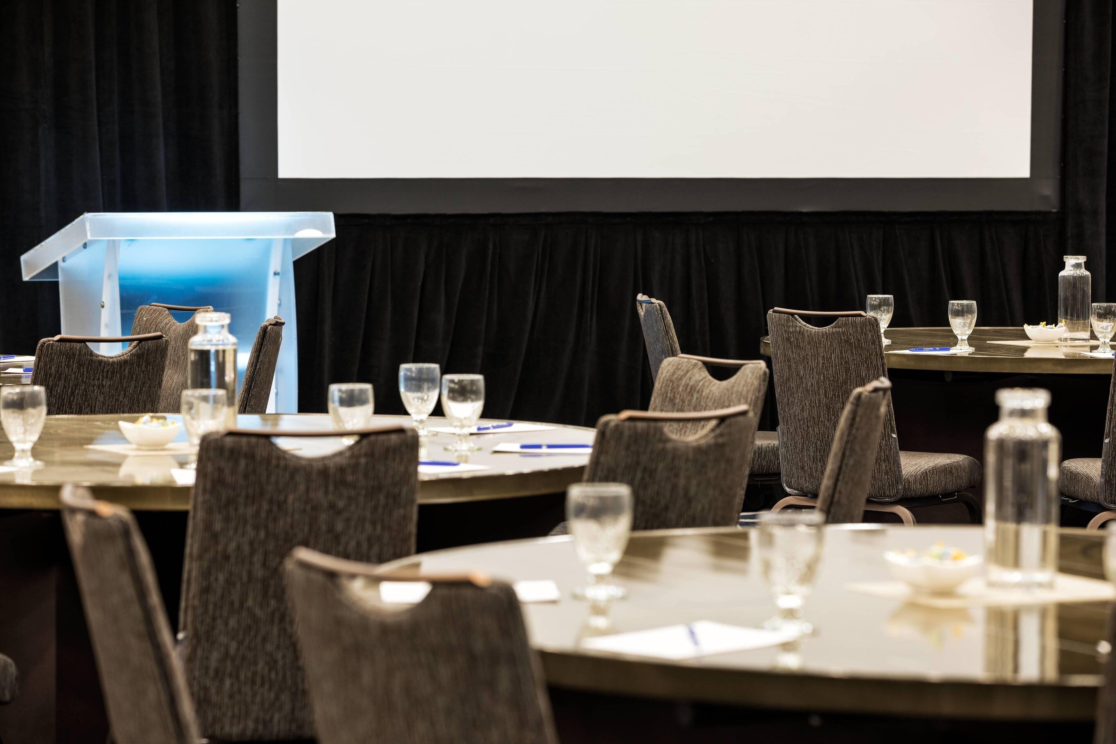A conference room with round tables, chairs, and a podium with a screen.