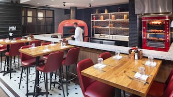 A restaurant with wooden tables, red chairs, a brick wall, and a pizza oven.