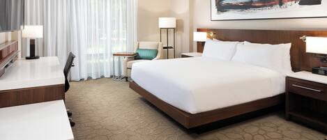 A hotel room with a large bed, a desk, a chair, a small table, and a lamp.