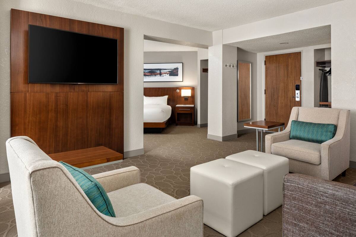 Habitación de hotel con televisor de pantalla plana montado en un panel de madera, cama con cabecera, zona de estar con sofá y reposapiés, y una mesita.