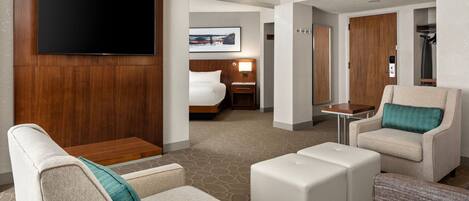 A hotel room with a flat-screen TV mounted on a wooden panel, a bed with a headboard, a seating area with a sofa and ottoman, and a small table.