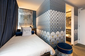 Quarto de hotel com cama, janela com cortinas, parede estampada e porta para o banheiro.