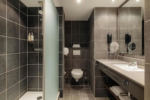 A modern bathroom with dark tiles, a shower, a toilet, a sink, and a mirror. - NYX Hotel Amsterdam Rembrandt Square (Amsterdam)