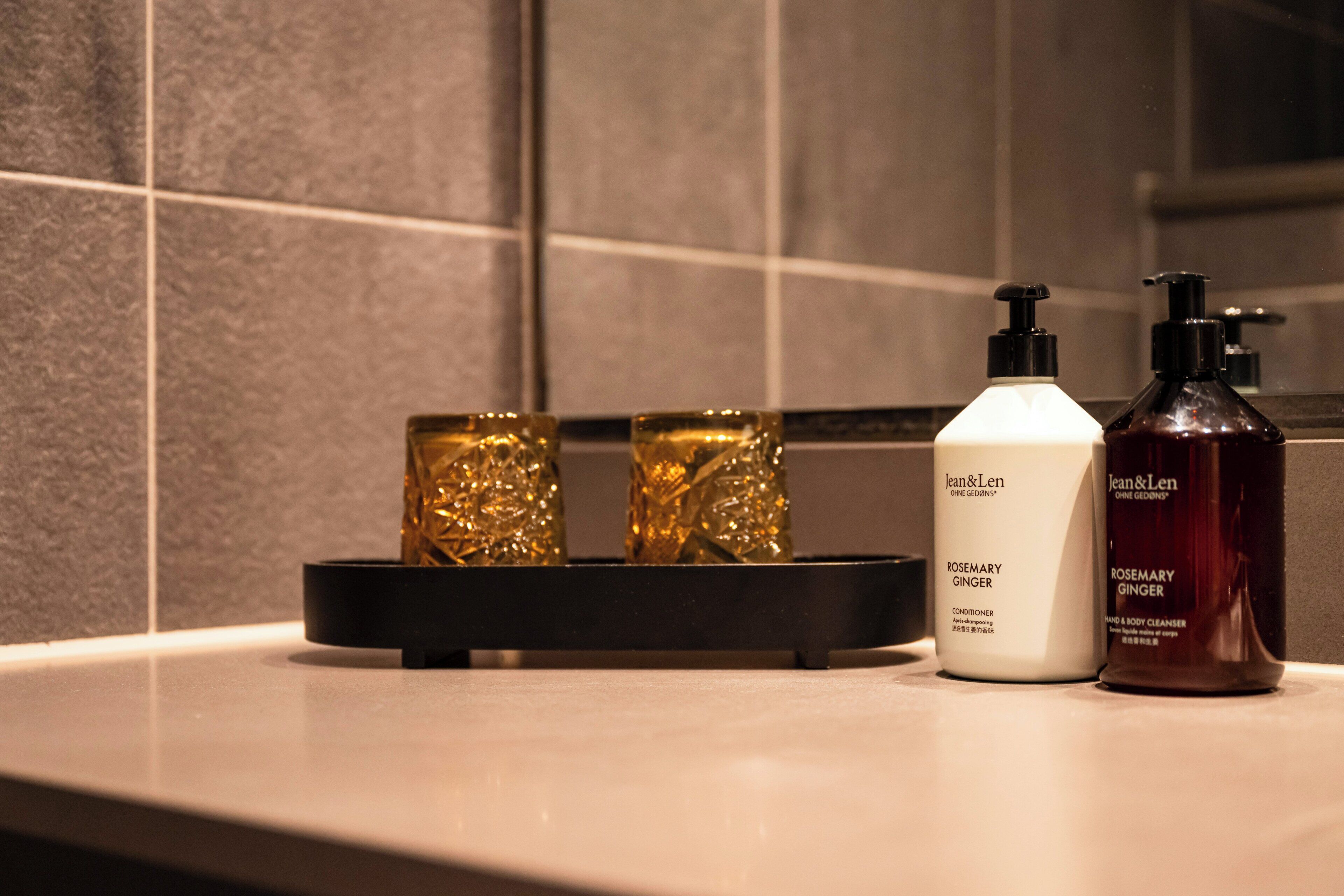 Una encimera de baño con dos botellas de limpiador corporal y de manos Romero y Jengibre Jean & Len, una botella blanca de acondicionador y tres botellas de vidrio dorado.