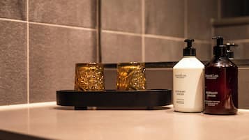 Una encimera de baño con dos botellas de limpiador corporal y de manos Romero y Jengibre Jean & Len, una botella blanca de acondicionador y tres botellas de vidrio dorado.