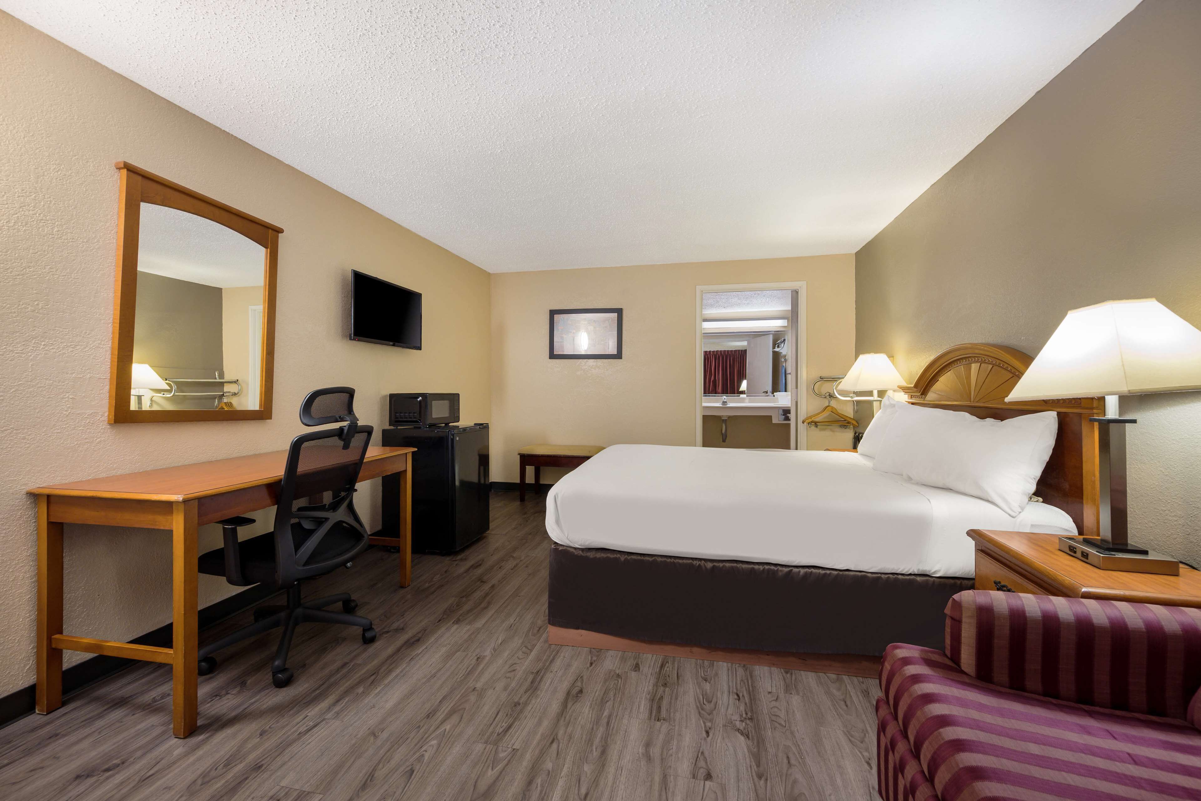 A hotel room with a bed, desk, chair, TV, and a small table with a lamp.