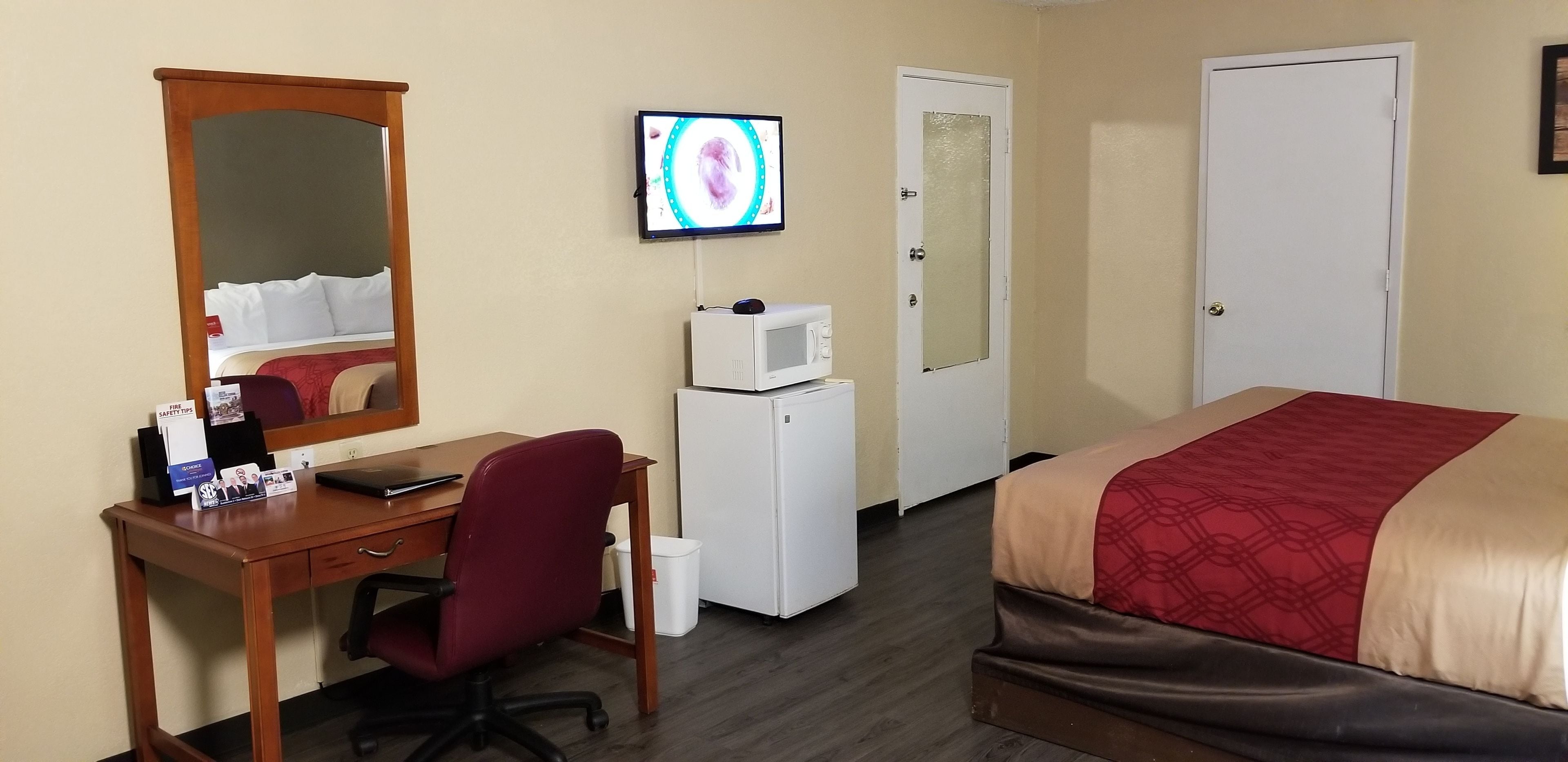Una habitación de hotel con una cama, un escritorio con computadora, una silla, un televisor, un microondas y una puerta.