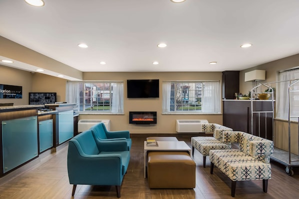 A modern hotel room with a kitchen, a fireplace, a flat-screen TV, and a seating area with a sofa and ottoman.