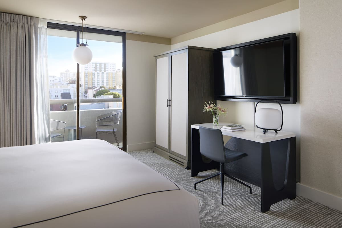 A hotel room with a bed, a desk, a chair, a TV, and a view of the city through a large window.