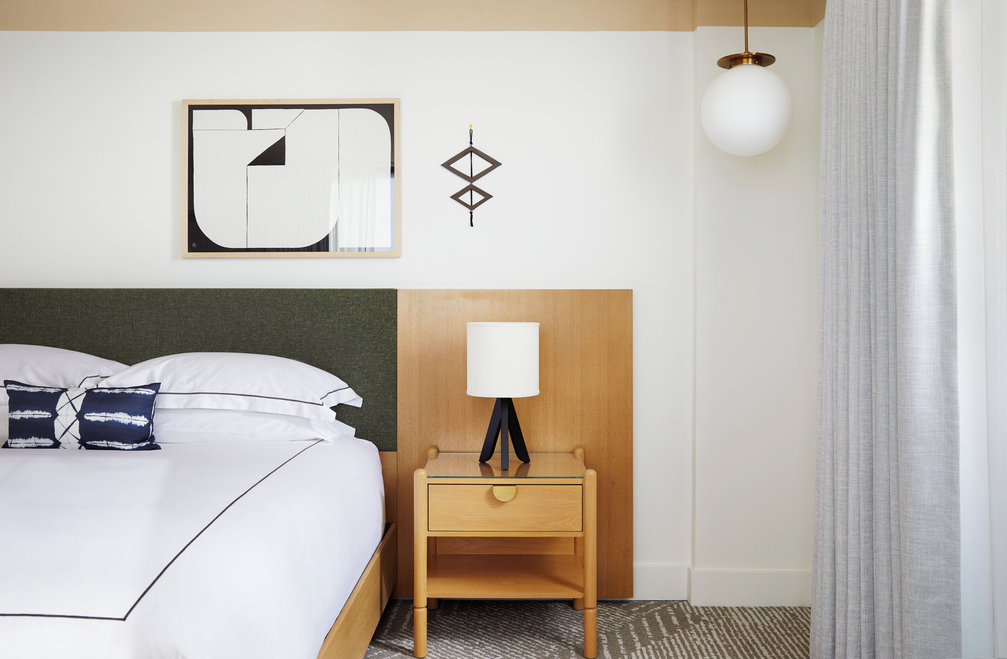 A hotel room with a bed, a nightstand with a lamp, a wall-mounted artwork, and a window with curtains.