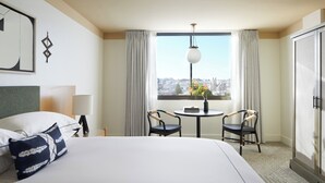 Una habitación de hotel con una cama, dos sillones, una mesa, una ventana con cortinas, una lámpara y vistas a la ciudad.