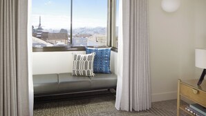 Una habitación con vistas a un paisaje urbano a través de un ventanal grande, un sofá con cojines estampados, una lámpara y una mesita de madera.