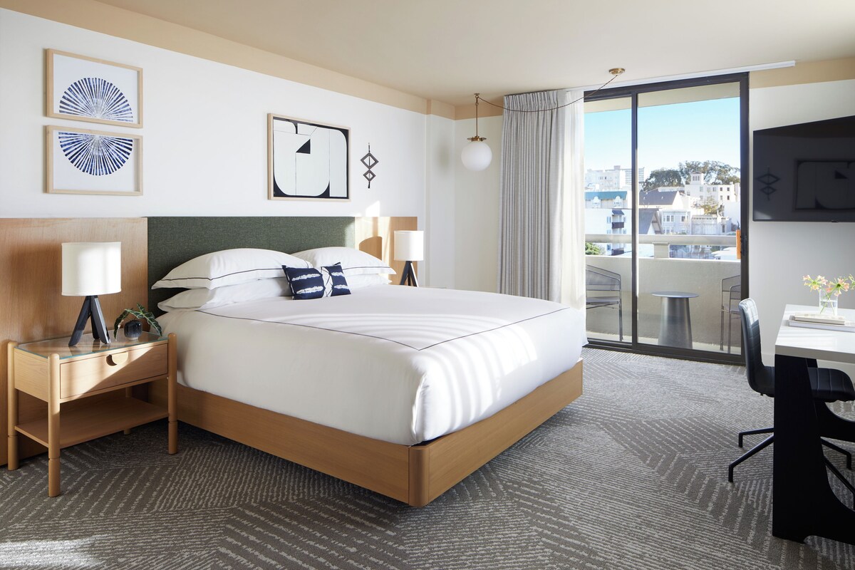 Habitación de hotel con cama, mesitas de noche, escritorio, silla, televisión y balcón con vistas.