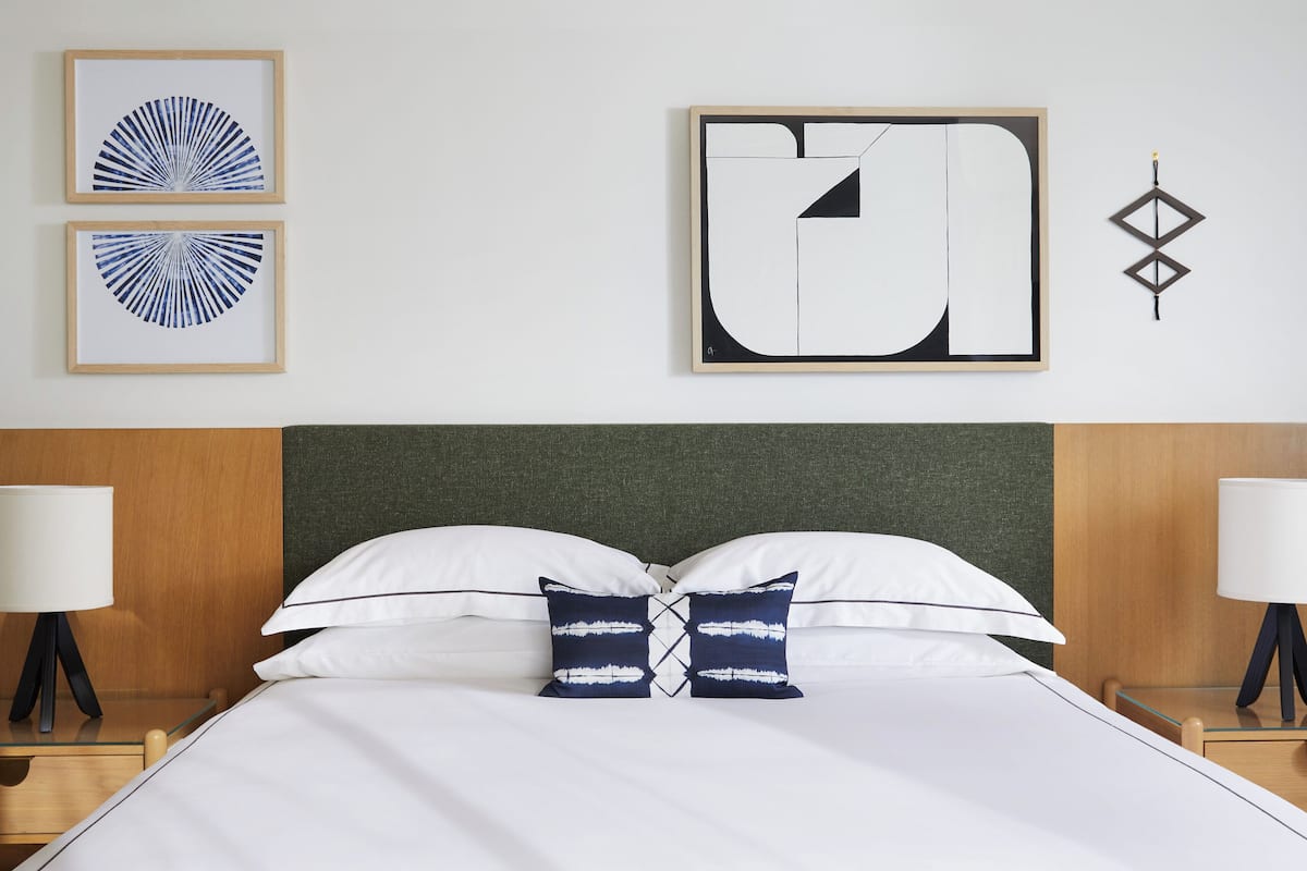A hotel room with a bed, two bedside tables, a nightstand, a wall-mounted lamp, and two framed artworks.
